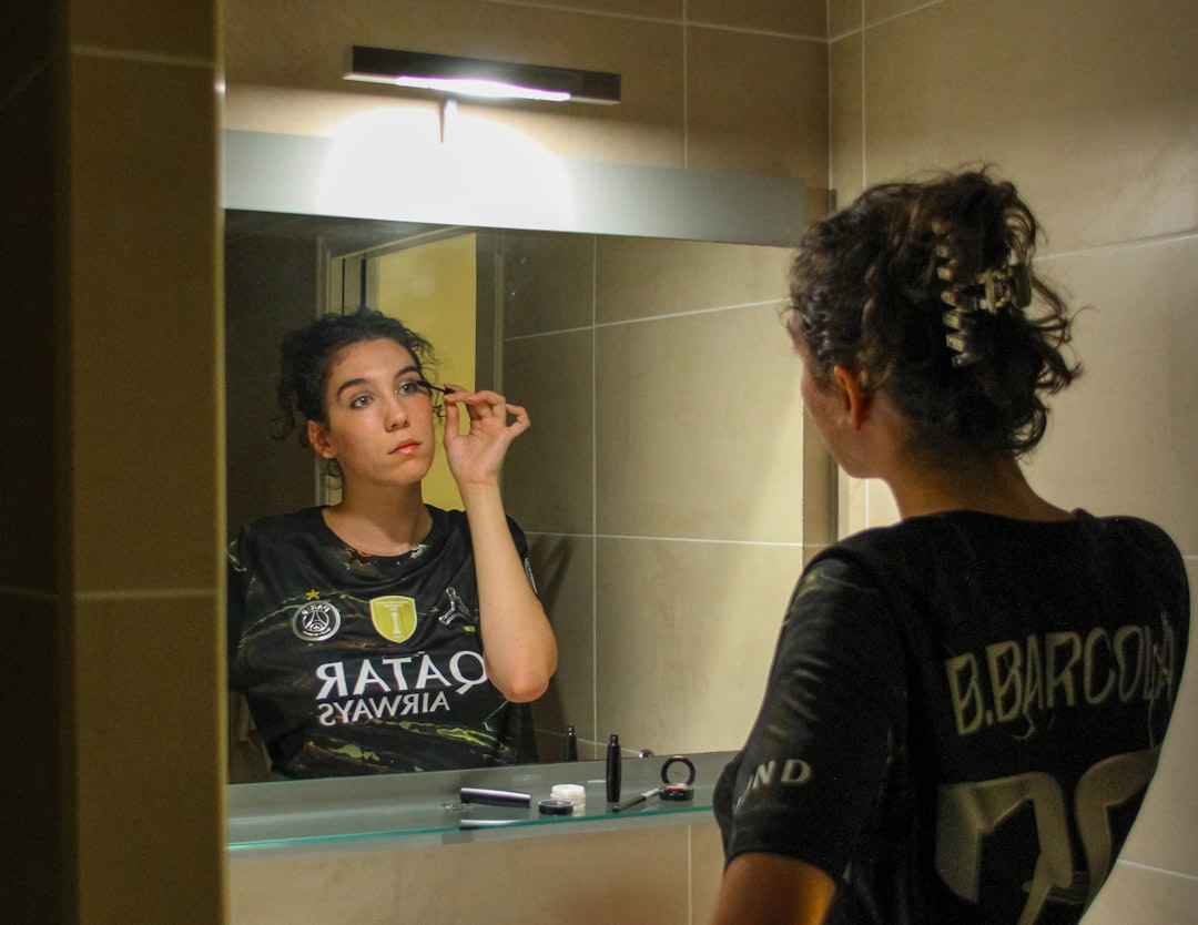 A woman applying mascara while looking into a bathroom mirror